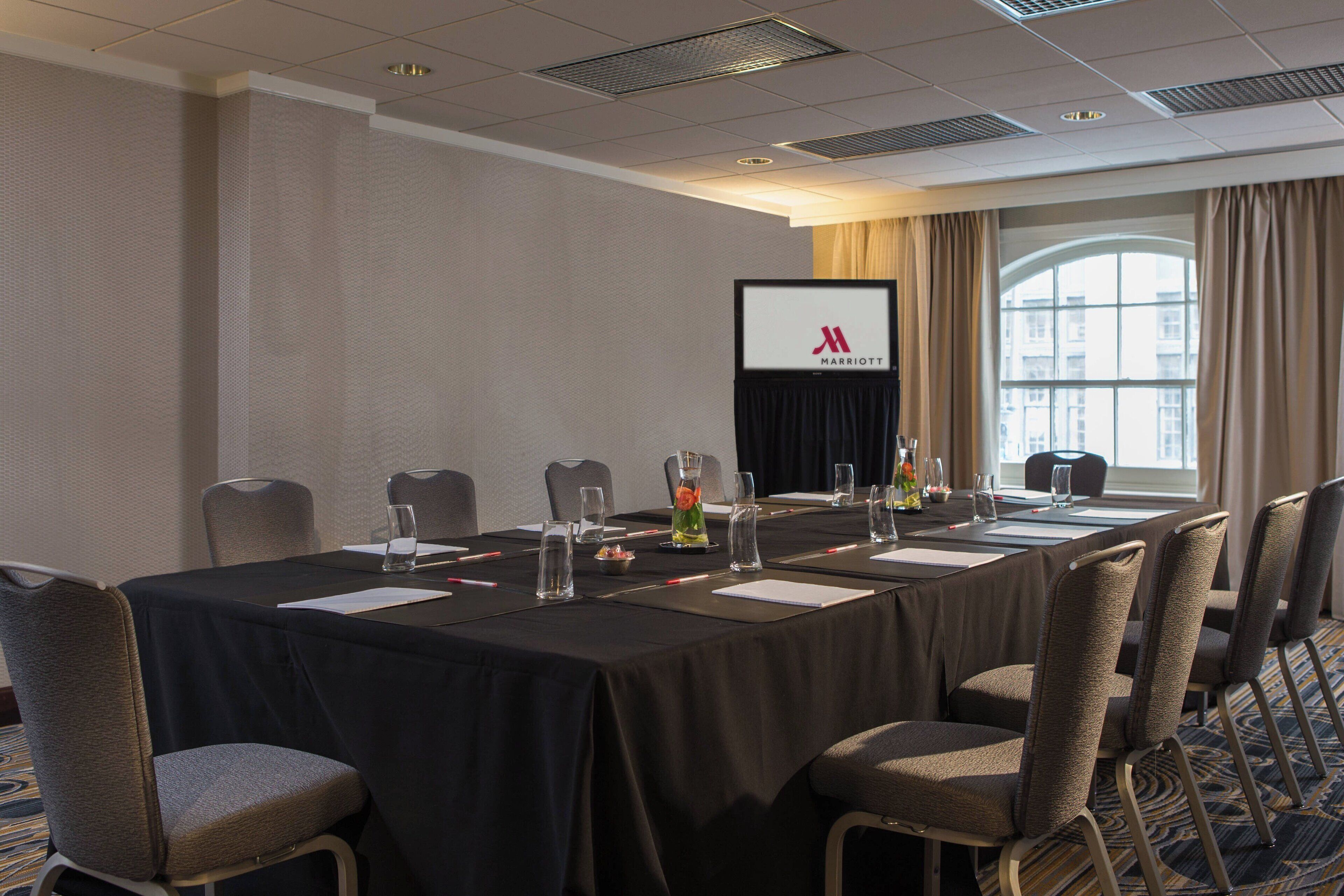 A conference room with a long table, chairs, a screen displaying the Marriott logo, and a window with curtains.