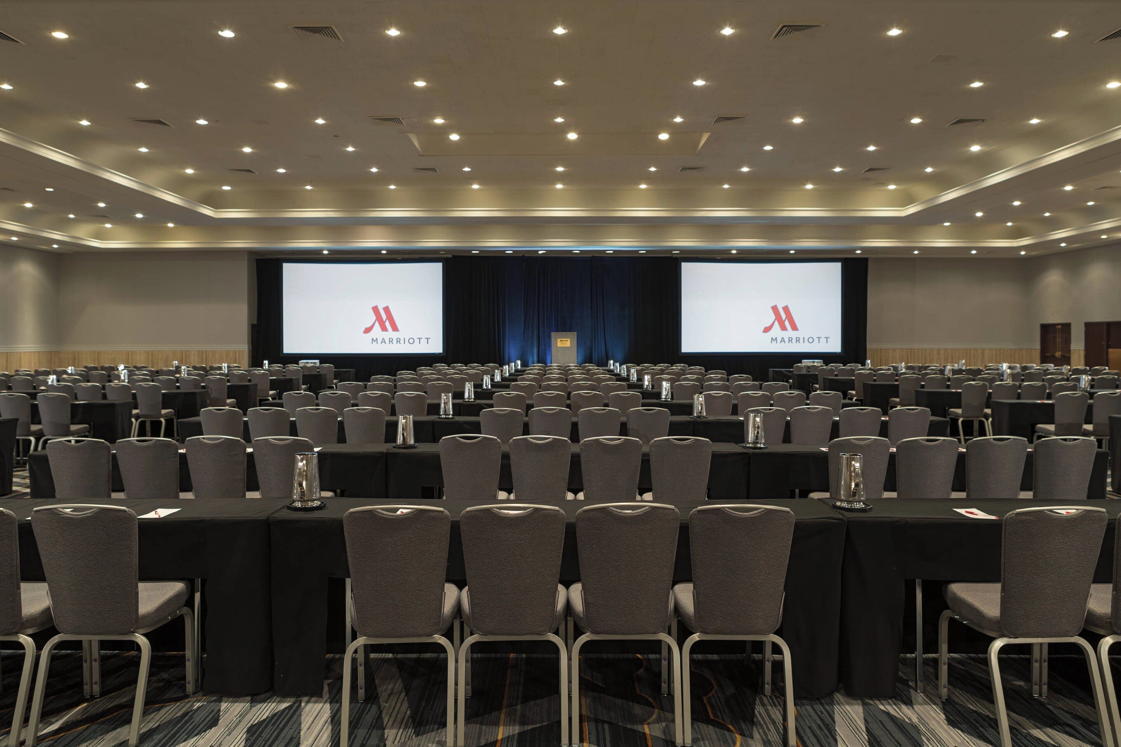 A large conference room with rows of tables and chairs, a stage with a screen displaying the Marriott logo, and a dark blue curtain backdrop.