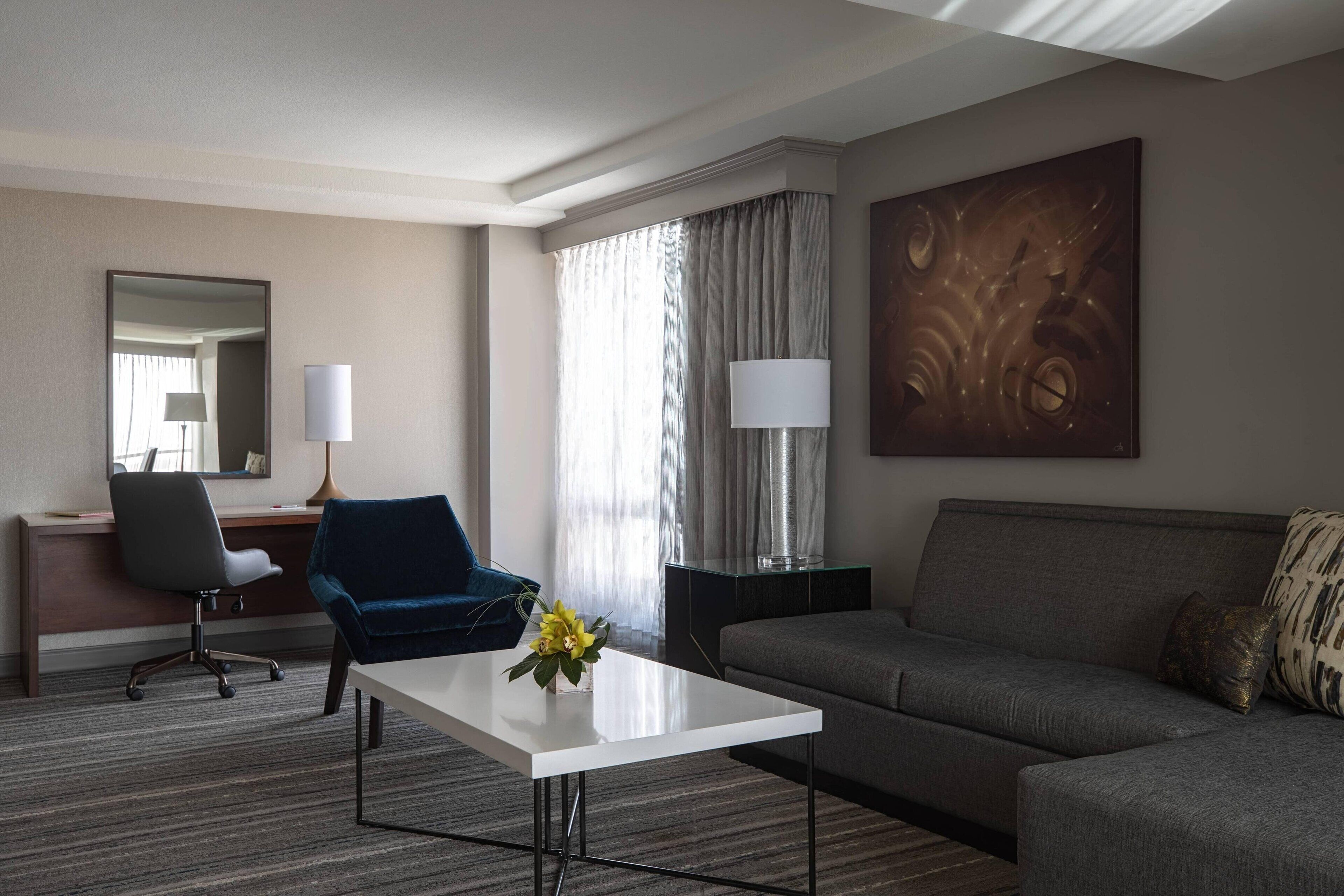Habitación de hotel con un sofá gris, un sillón azul, una mesa de centro blanca con un jarrón de flores, un escritorio con una silla, una lámpara y un cuadro abstracto grande en la pared.