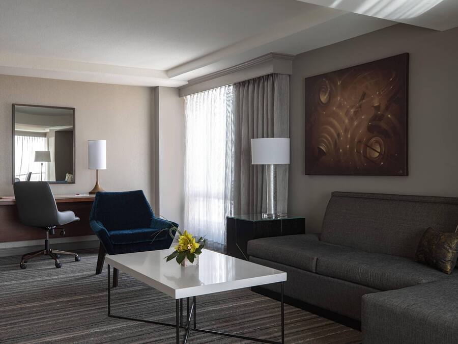 A hotel room with a grey sofa, a blue armchair, a white coffee table with a vase of flowers, a desk with a chair, a lamp, and a large abstract painting on the wall.