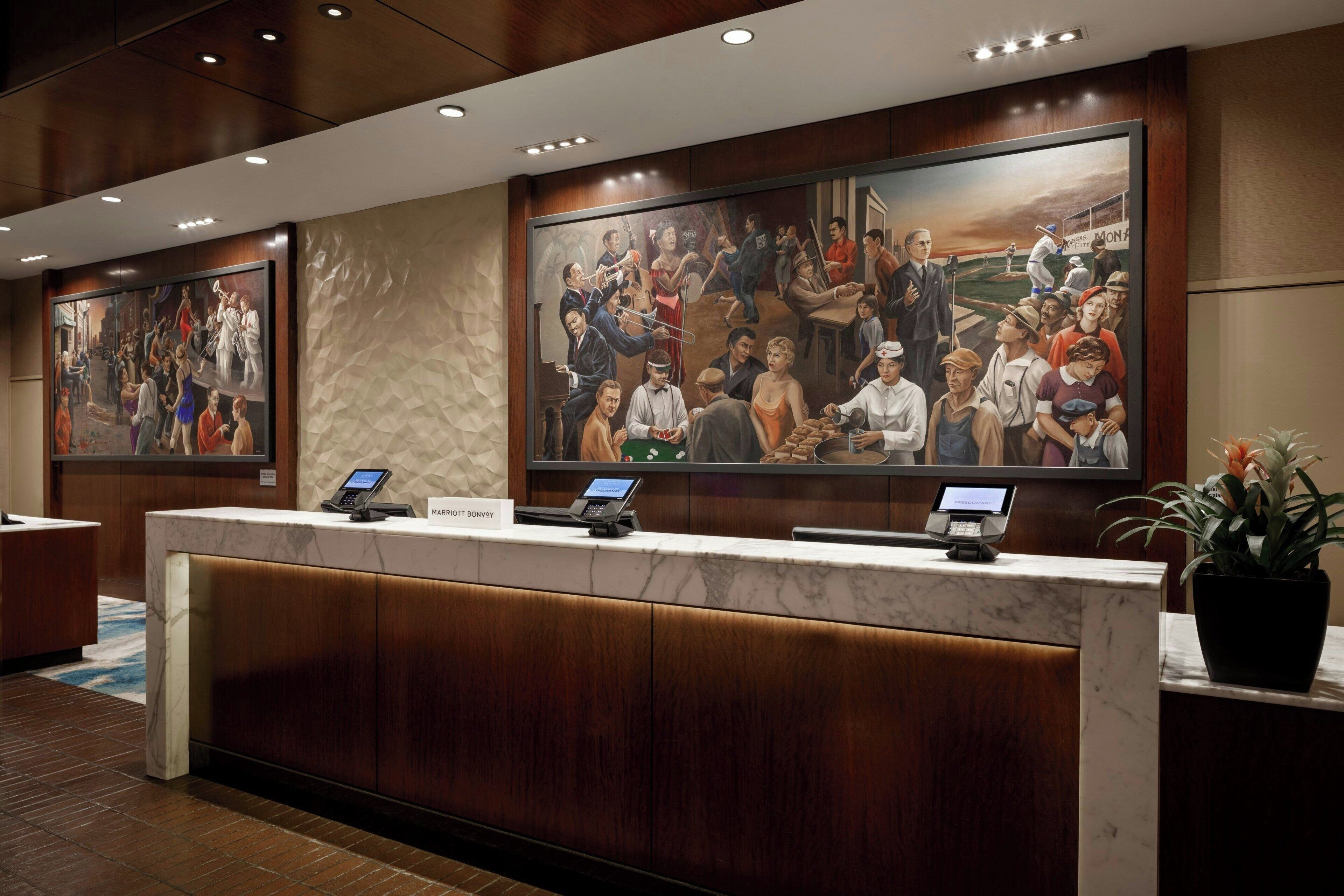 Un mostrador de recepción de hotel con encimera de mármol, dos quioscos digitales y un mural grande que representa una escena histórica.