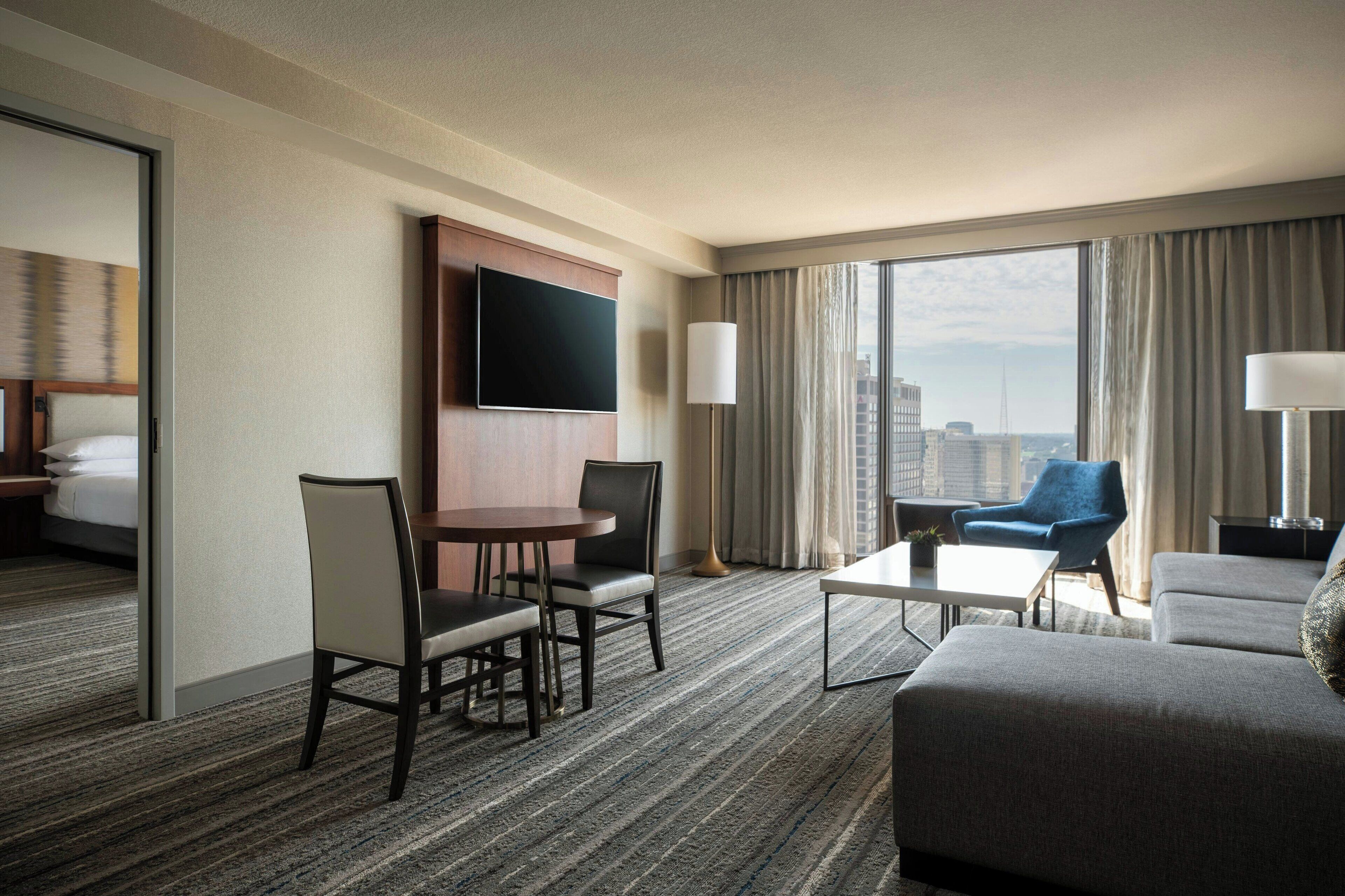 A hotel room with a large bed, a TV mounted on the wall, a dining area with chairs and a table, a sofa, and a view of the city through large windows.