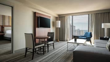 A hotel room with a large bed, a TV mounted on the wall, a dining area with chairs and a table, a sofa, and a view of the city through large windows.