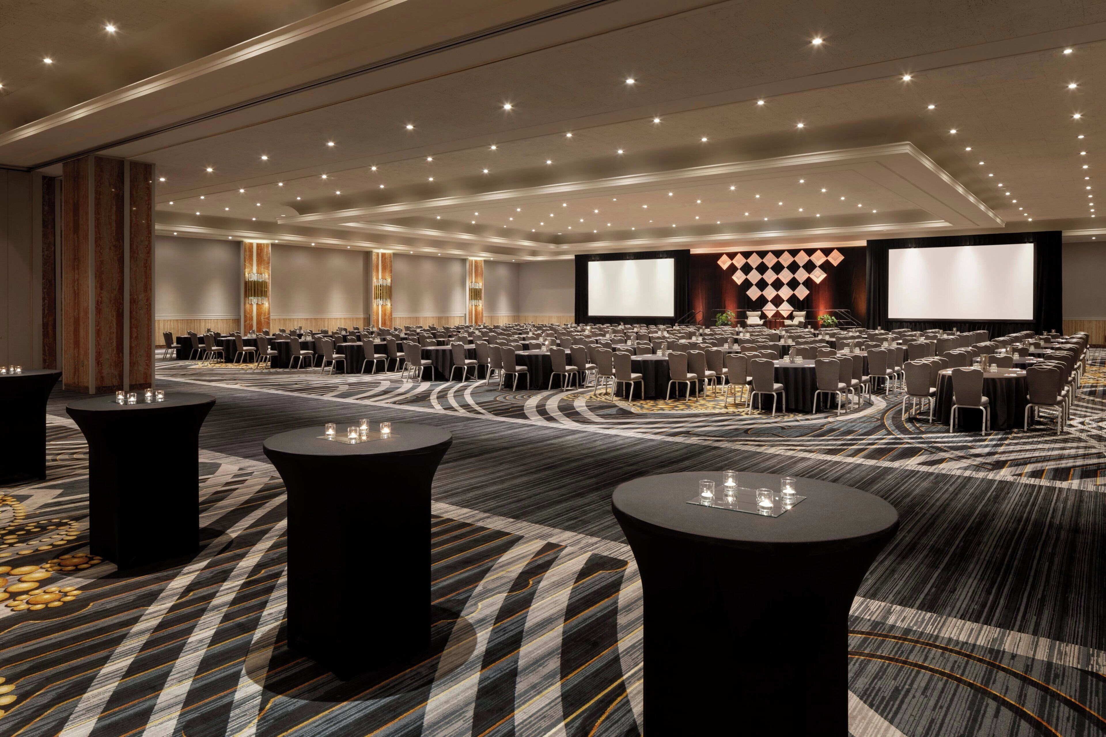 A large conference room with rows of tables and chairs, a stage with a large screen, and a patterned carpet.