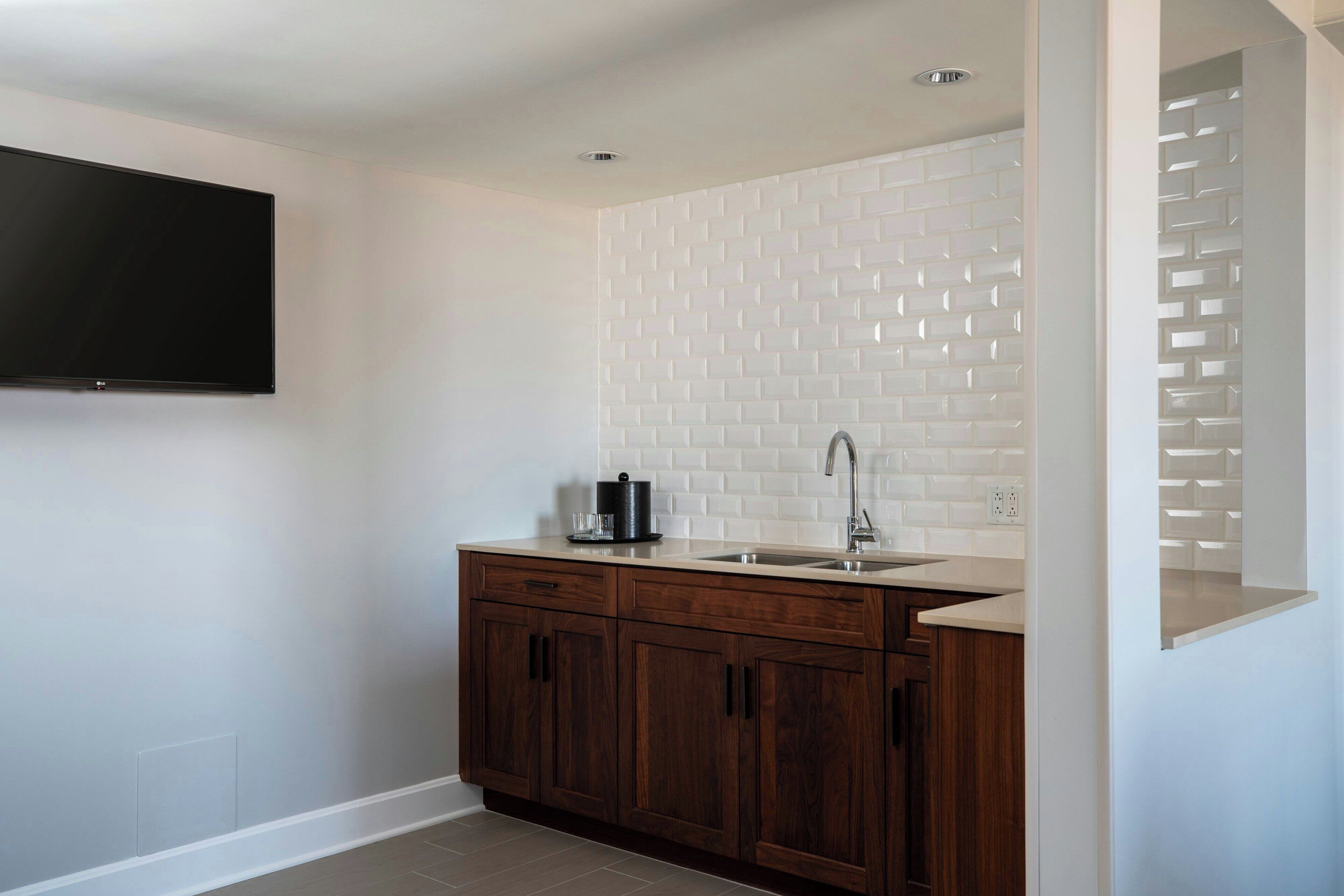 A modern kitchen with a white tiled backsplash, wooden cabinets, a stainless steel sink, and a flat-screen TV mounted on the wall.