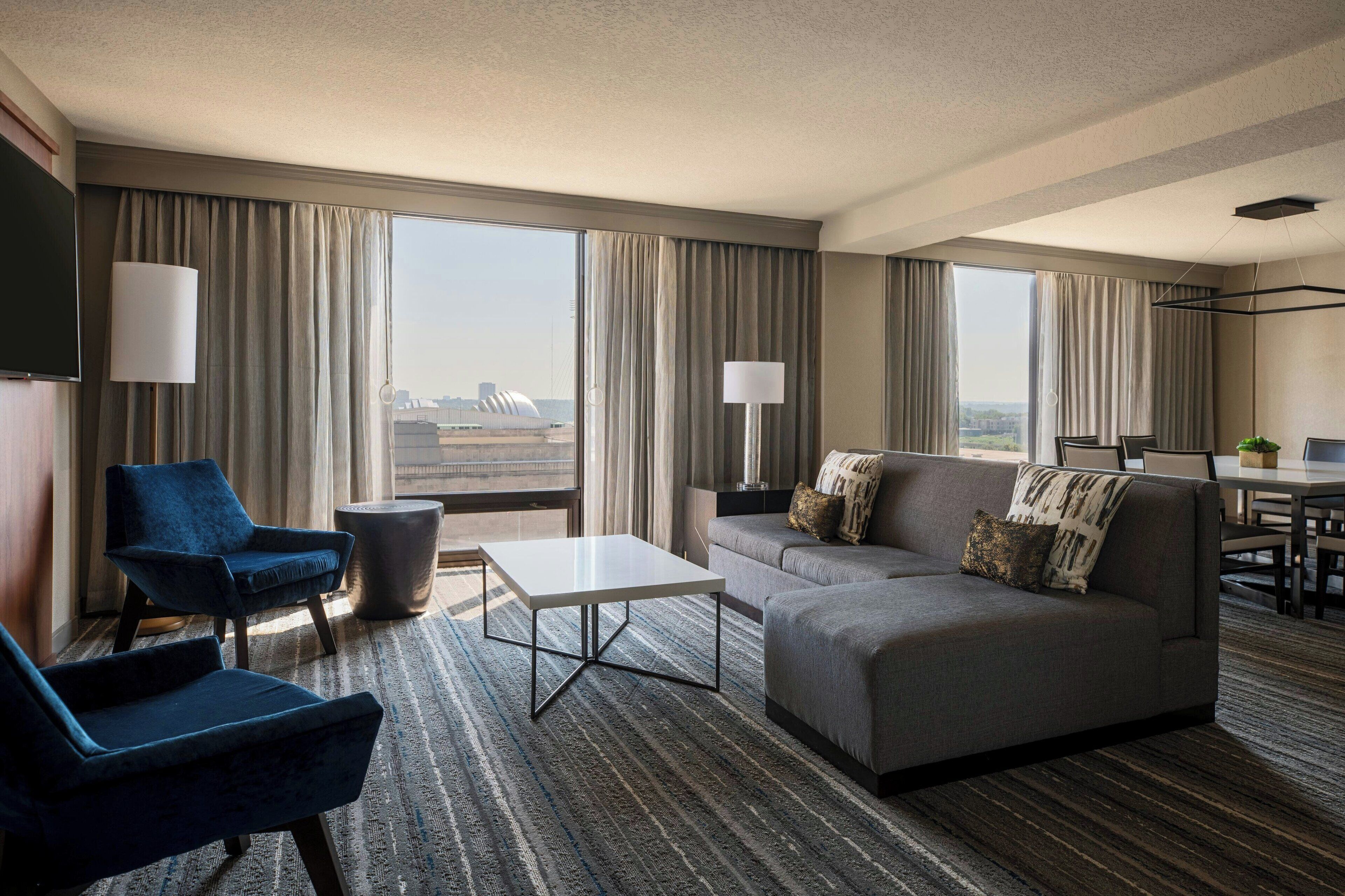 A modern hotel room with a sofa, two armchairs, a coffee table, and a TV. Large windows offer a view of the cityscape.