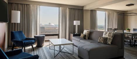 A modern hotel room with a sofa, two armchairs, a coffee table, and a TV. Large windows offer a view of the cityscape.