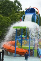 Un área de juegos acuáticos con un tobogán y chorros de agua, rodeada de follaje.