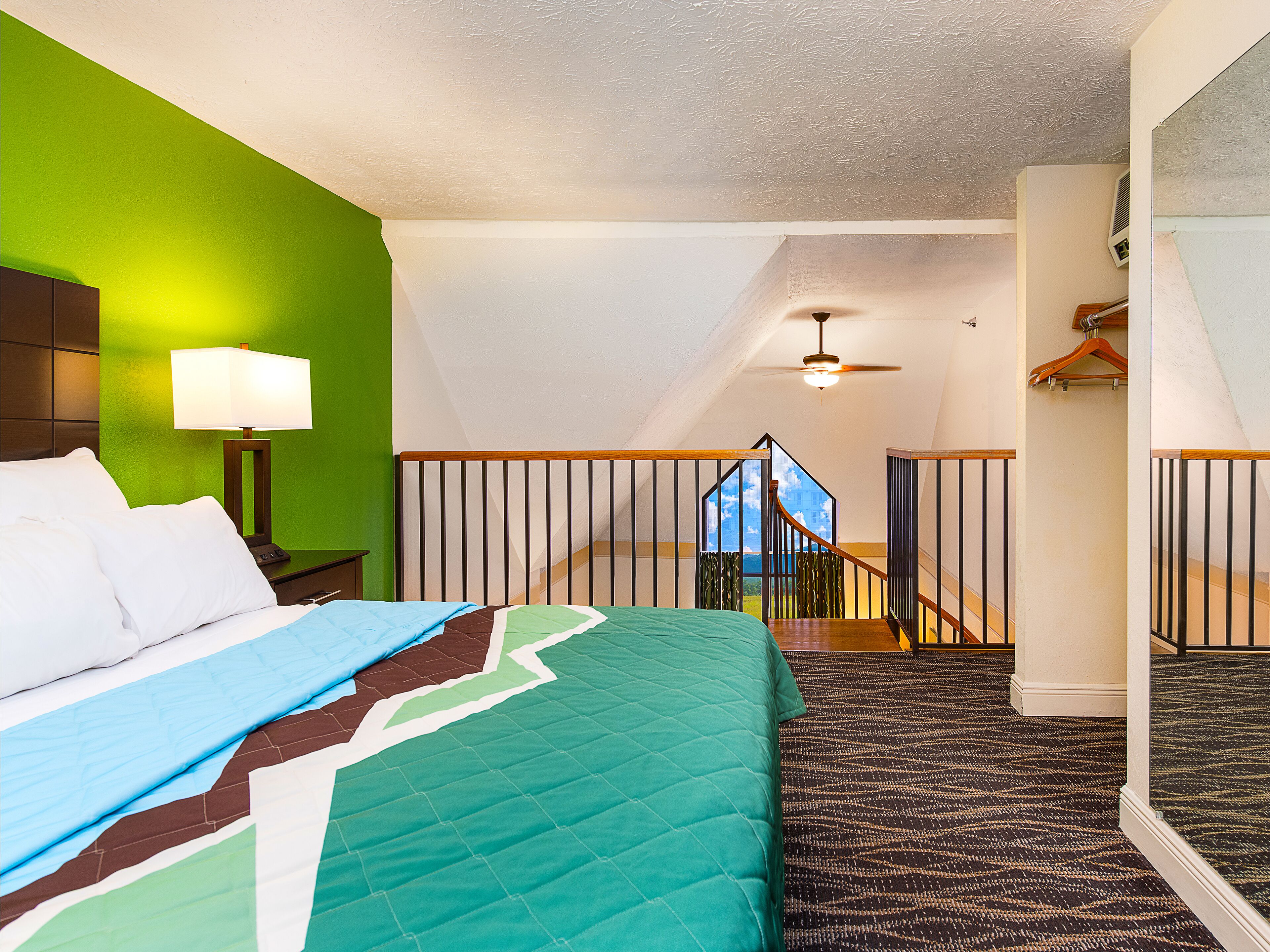 A hotel room with a bed, a nightstand, a lamp, a view of the outdoors, and a staircase.