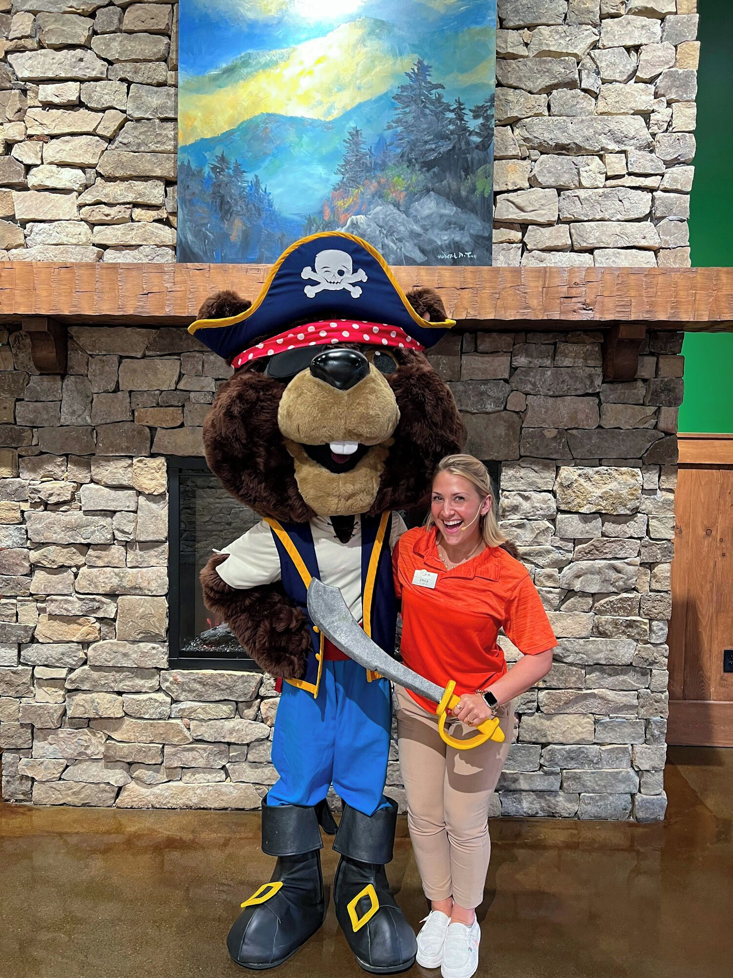 Une mascotte déguisée en ours, portant un chapeau de pirate et tenant une épée, se tient devant un mur de pierre sur lequel est accrochée une peinture.