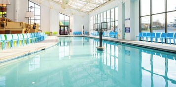 An indoor swimming pool with clear water, blue chairs along the sides, and large windows.