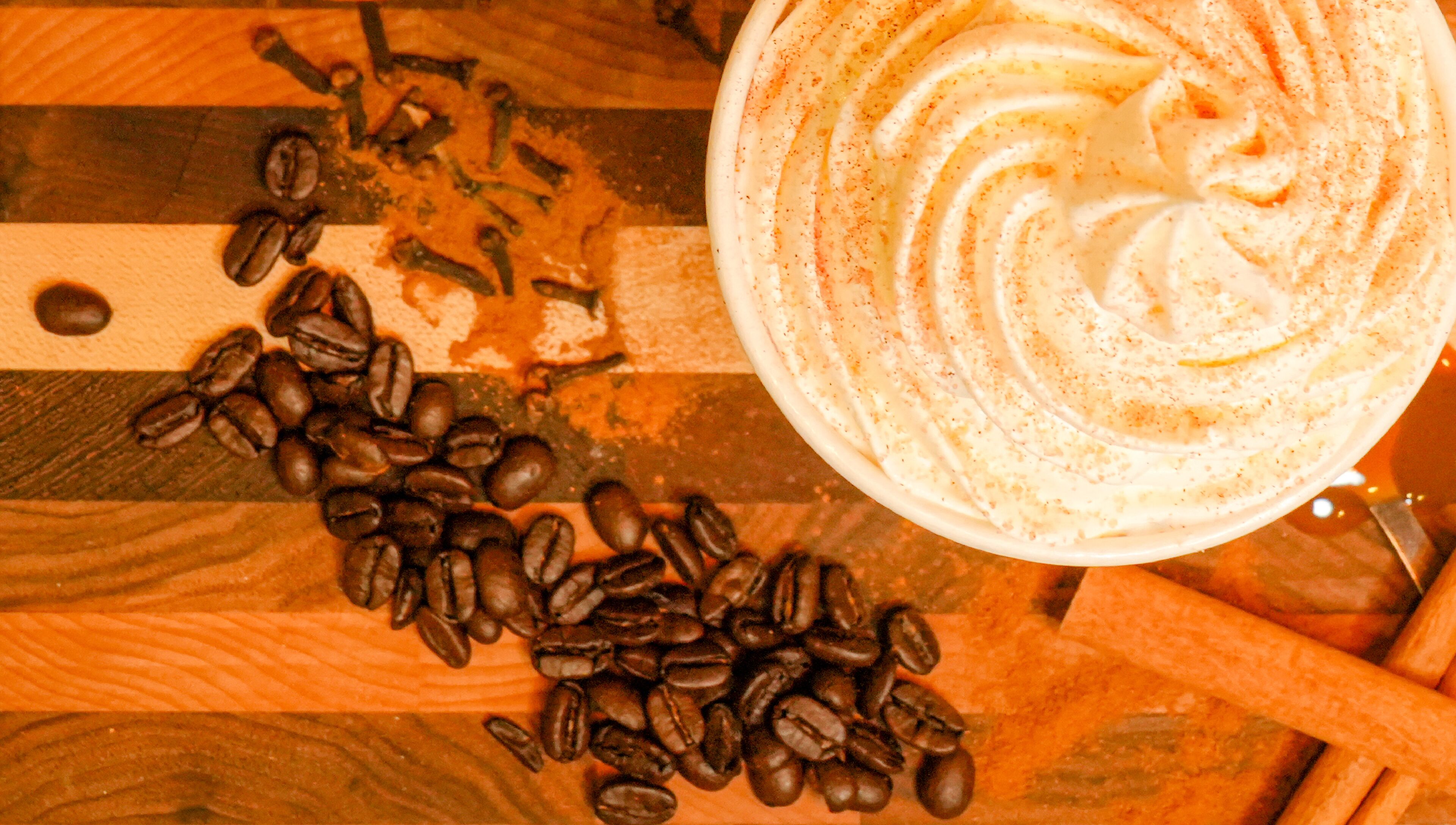 A close-up of a coffee drink with whipped cream and cinnamon, surrounded by coffee beans and a cinnamon stick on a wooden surface.