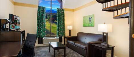 A hotel room with a large window, a desk with a computer, a sofa, a coffee table, and a lamp.