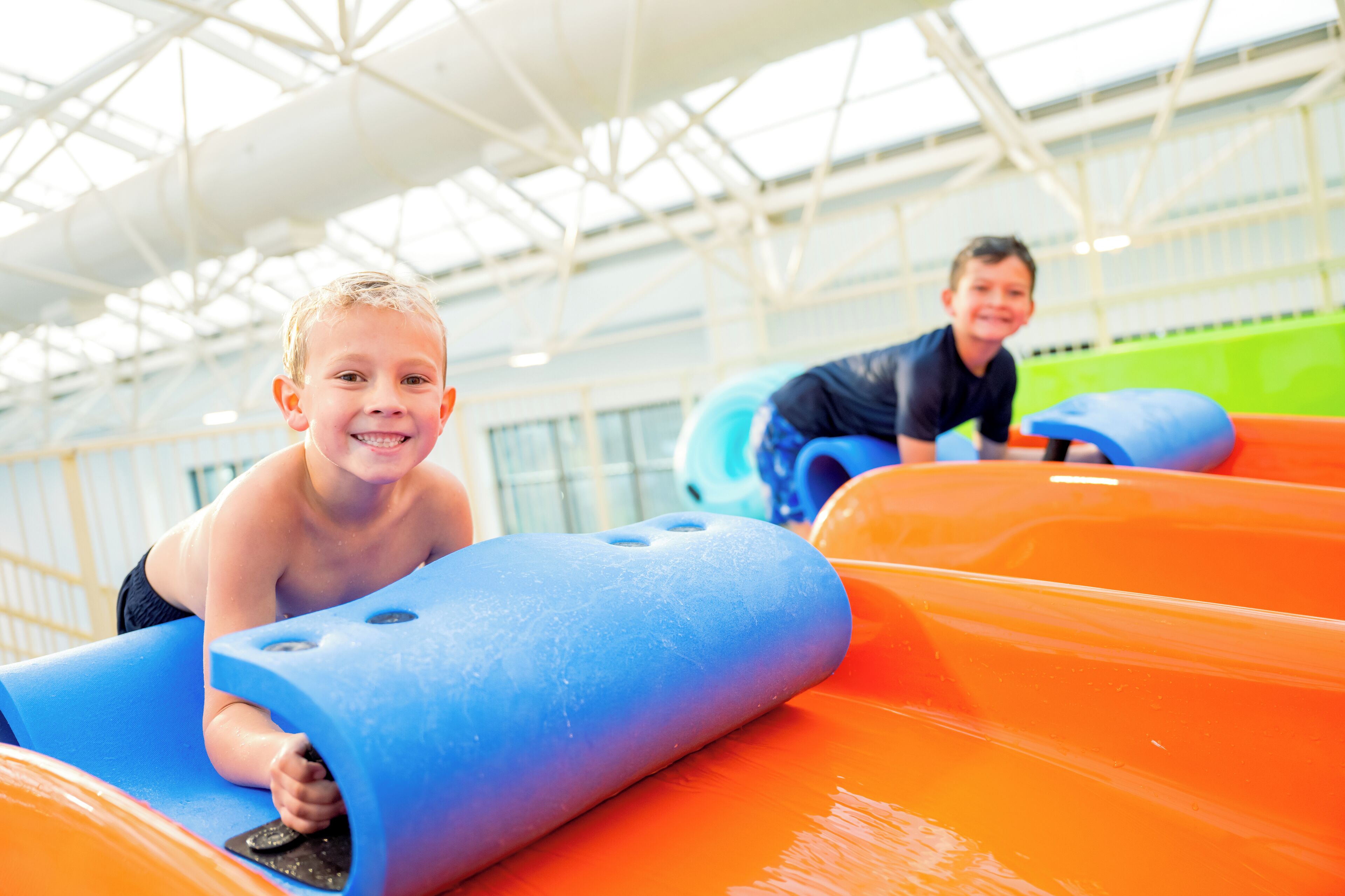 Children playing on a water slide in an indoor facility with a high ceiling and large windows.
