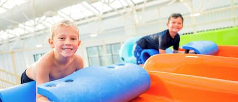 Children playing on a water slide in an indoor facility with a high ceiling and large windows.