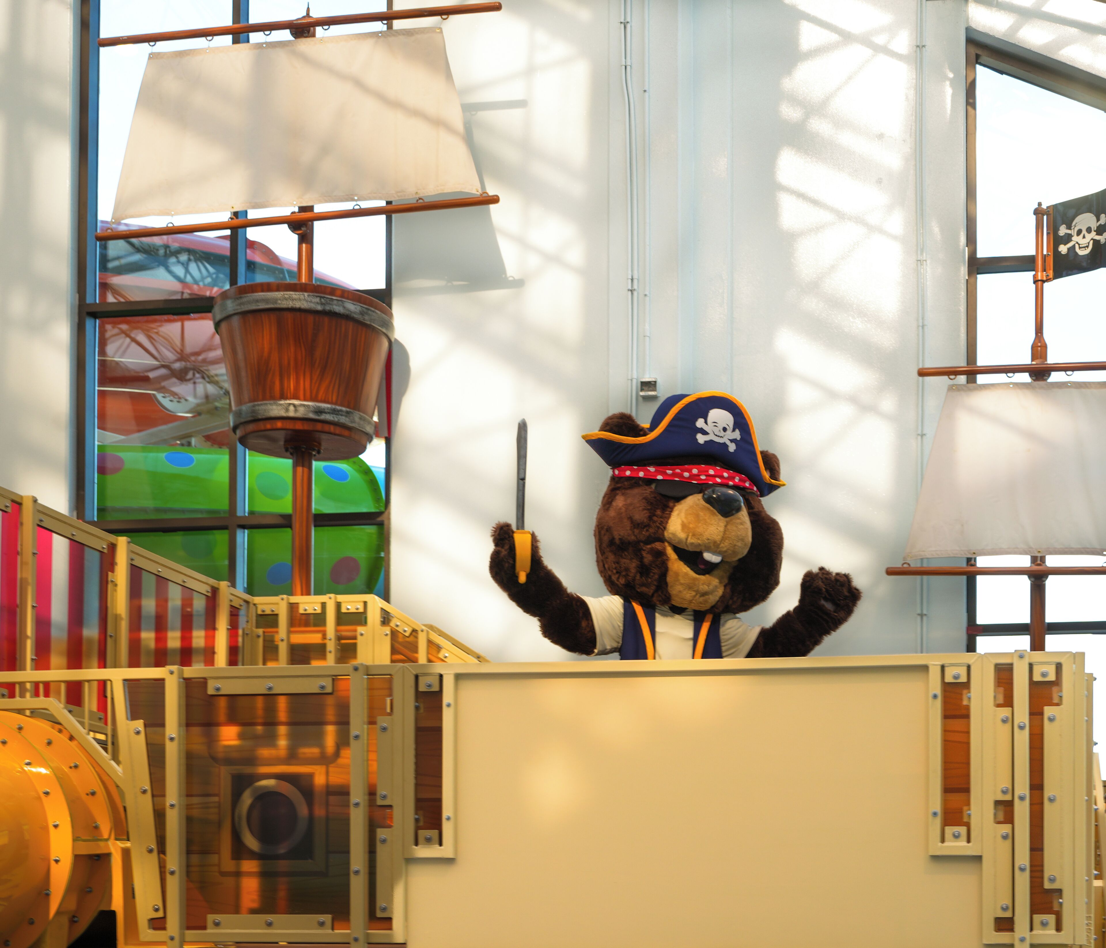 A mascot in a pirate costume standing in a room with large windows and a ship's wheel.