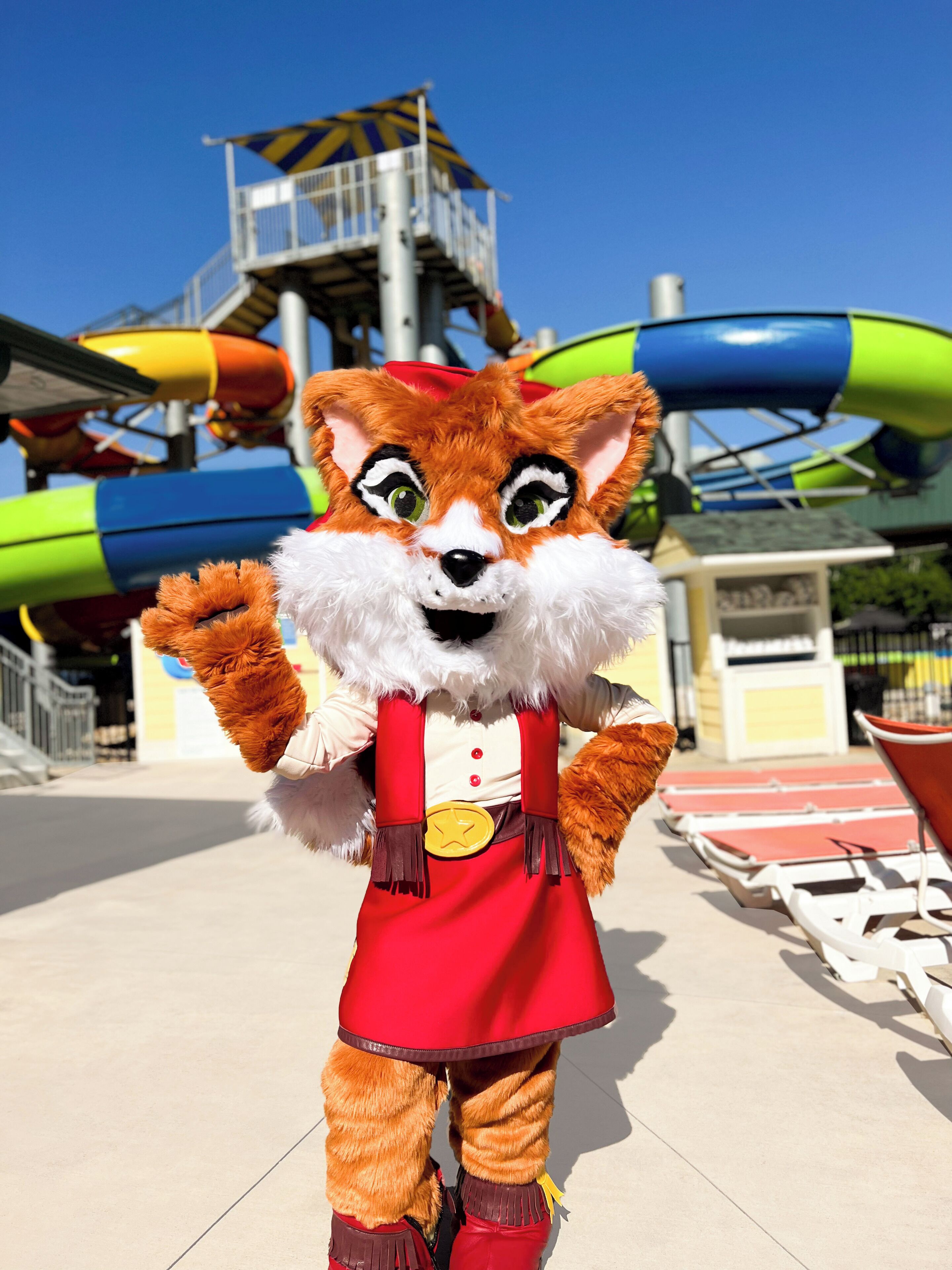 A mascot in a red and white outfit standing in front of a water park with slides and a lifeguard tower.