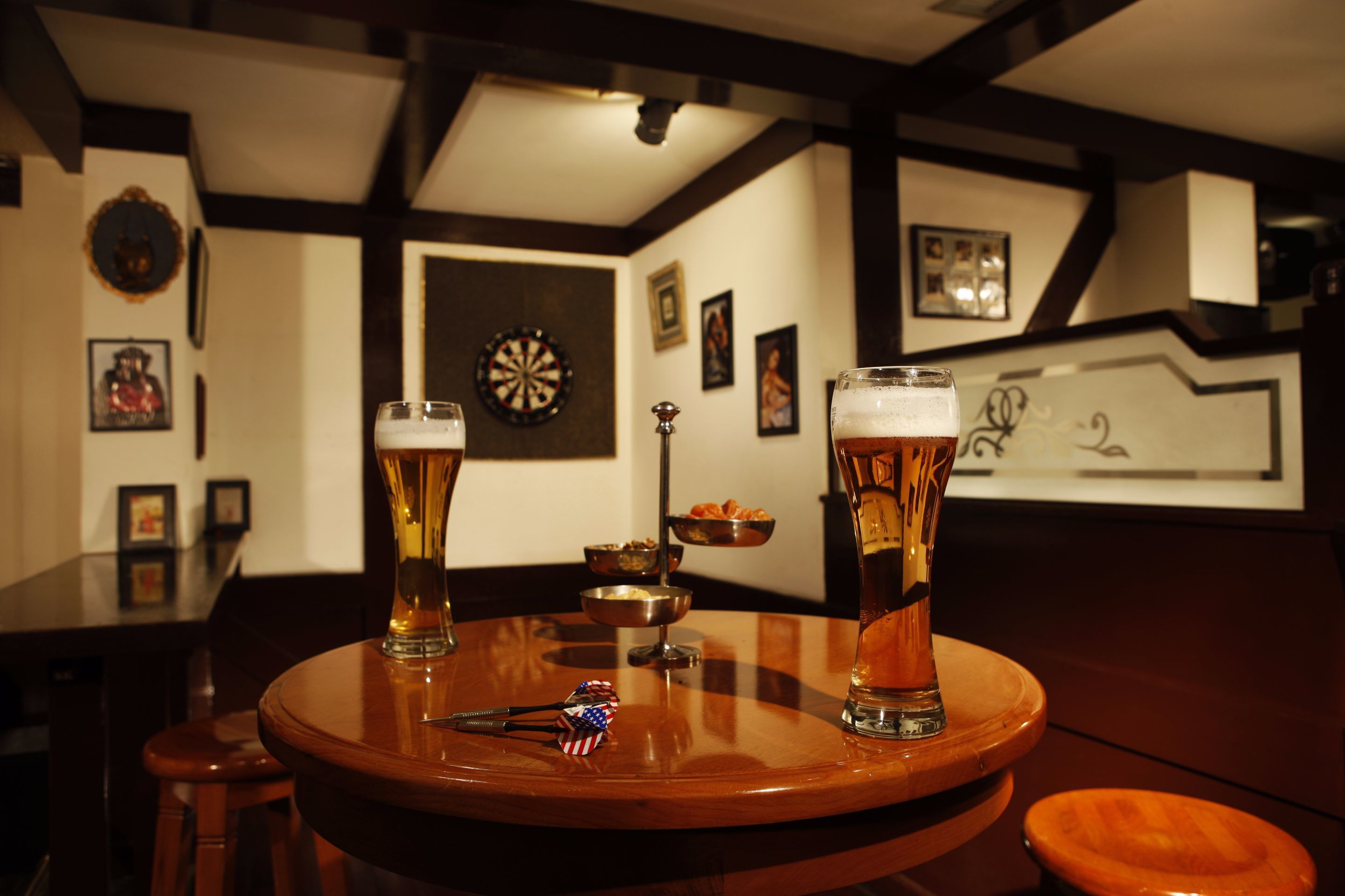 A bar setting with two glasses of beer, a dartboard, and a small metal bowl on a wooden table.