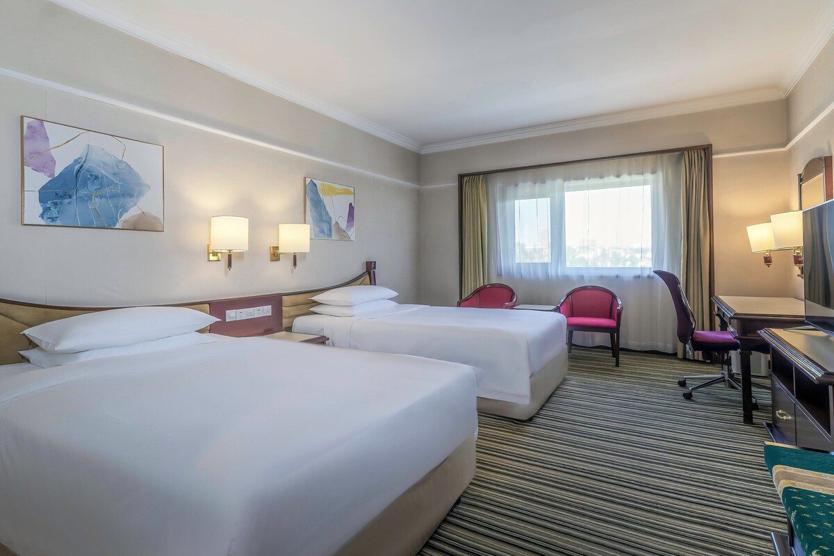 Habitación de hotel con dos camas, un escritorio, una silla y obras de arte en las paredes.