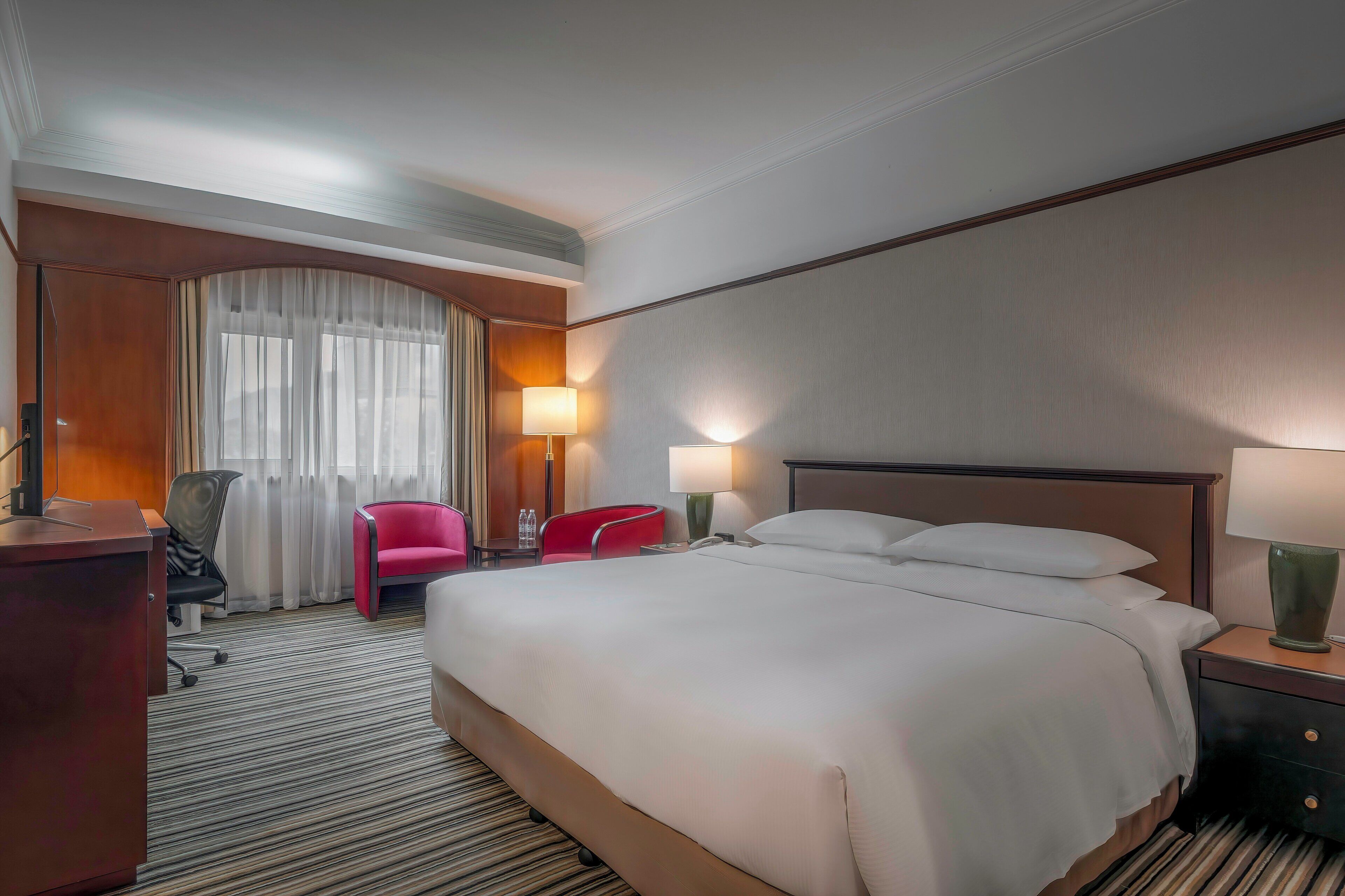 Une chambre d’hôtel avec un grand lit, un bureau, une chaise, une lampe et une fenêtre avec des rideaux.