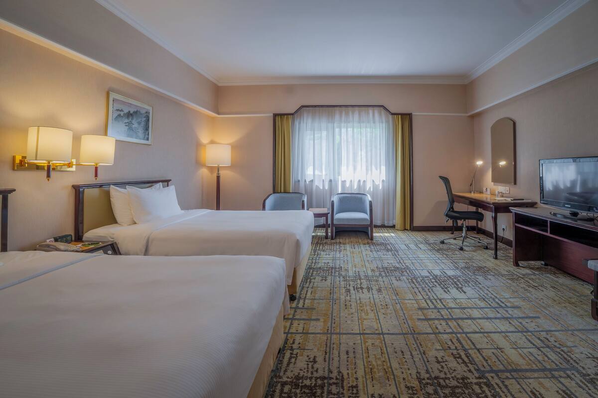 A hotel room with two beds, a desk, a chair, a television, and a large window with curtains.
