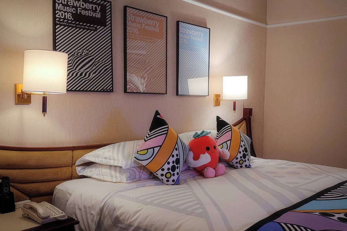 A hotel room with a bed, pillows, a lamp, a phone, and framed posters on the wall.