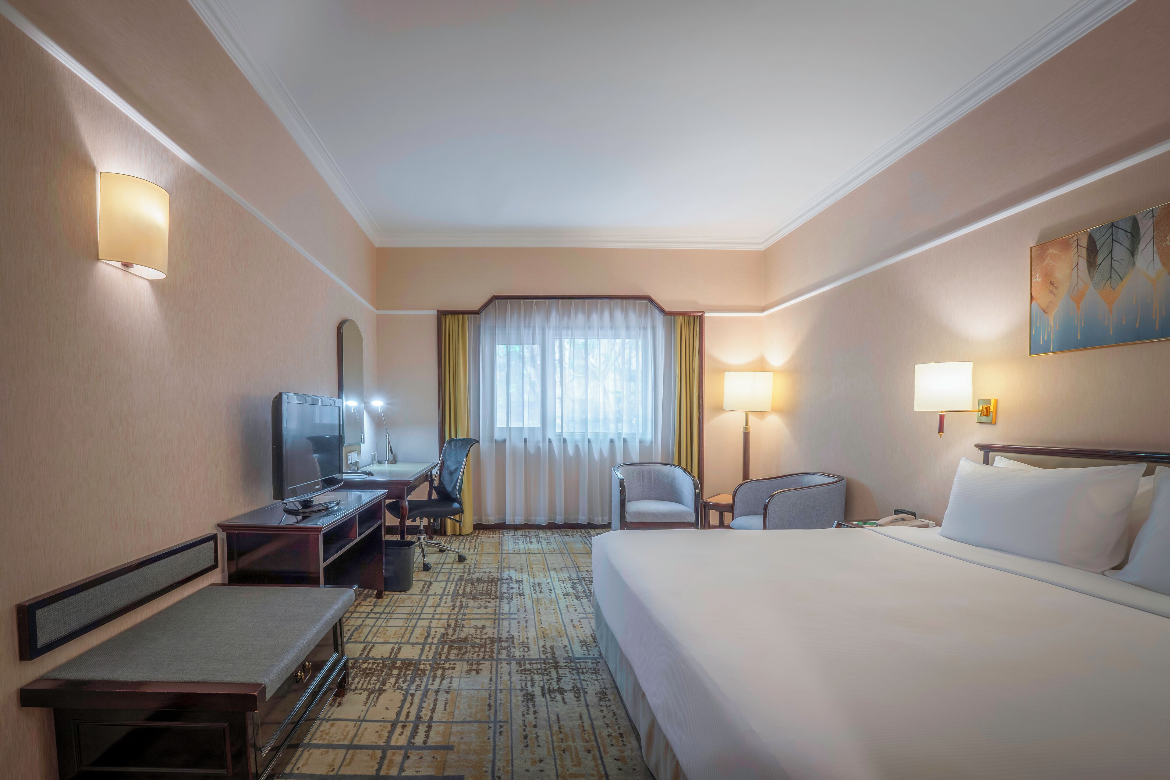 A hotel room with a bed, desk, chairs, a TV, and a window with curtains.