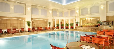 A large indoor swimming pool with red chairs and tables, surrounded by columns and potted plants.