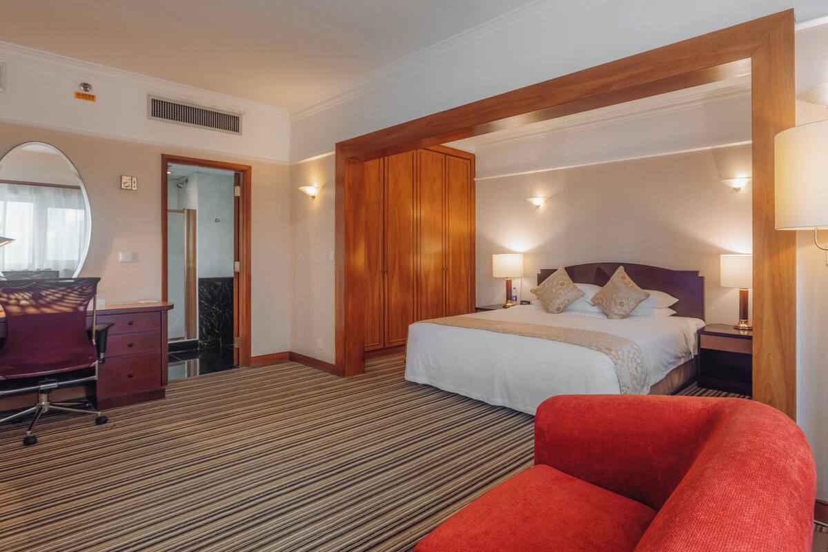 A hotel room with a large bed, a desk with a chair, a wardrobe, and a mirror.