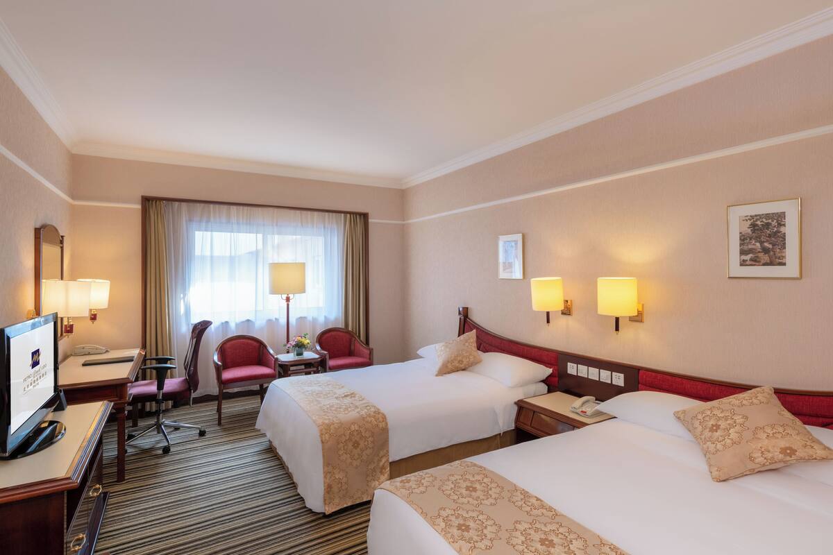 A hotel room with two beds, a desk with a computer, a chair, a TV, and a window with curtains.