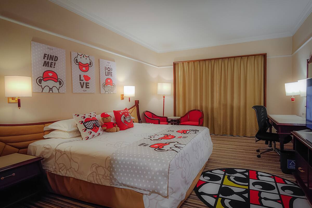 A hotel room with a bed, a desk, a chair, a TV, and artwork on the walls.