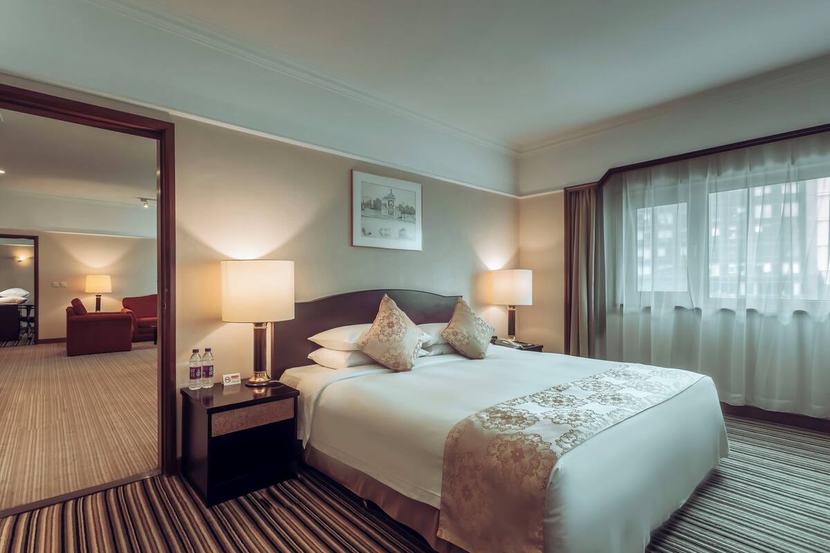 A hotel room with a large bed, two bedside lamps, a nightstand, a mirror, a red sofa, and a window with sheer curtains.