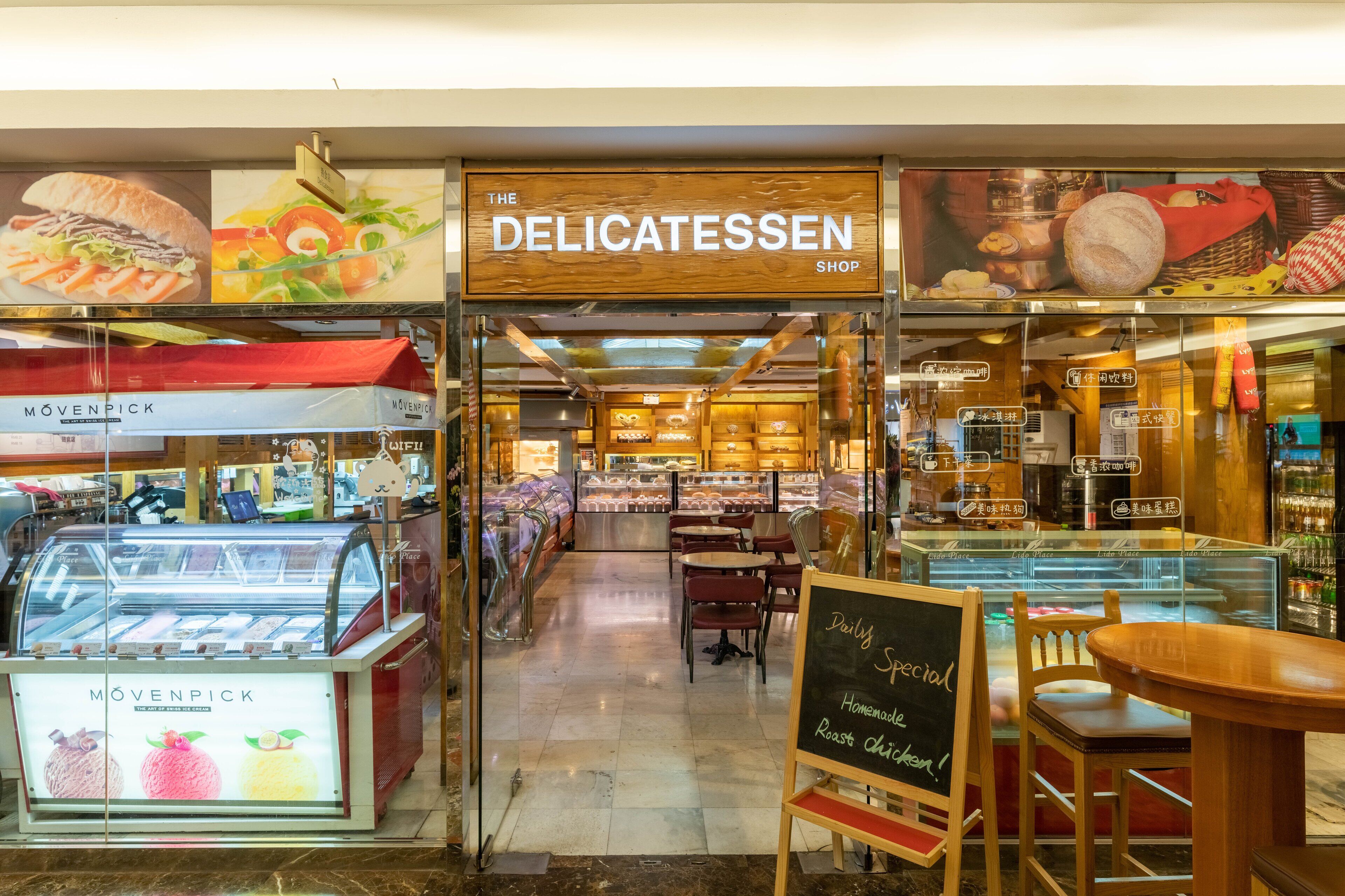 Una tienda de comida llamada 'The Delicatessen' con una exhibición de diversos productos alimenticios y una pizarra anunciando 'Pollo Asado Especial del Día'.