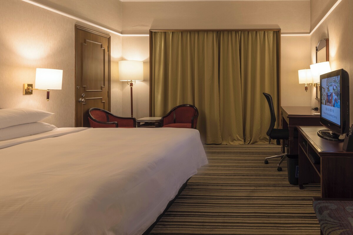 Habitación de hotel con una cama grande, dos sillas, un escritorio con televisor y una ventana con cortinas.