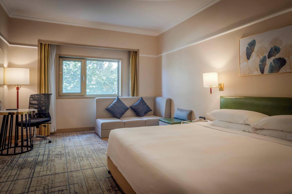 A hotel room with a large bed, a sofa, a desk with a chair, a window with curtains, and a wall-mounted lamp.