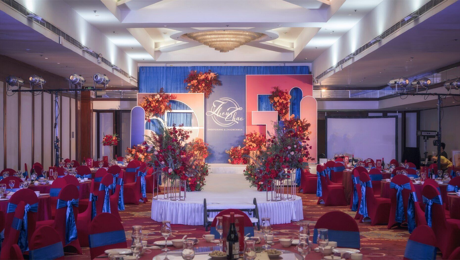 A banquet hall with round tables, red chairs, and blue sashes.