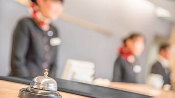 A hotel reception area with a wooden counter, a silver bell, and blurred figures of staff in the background.