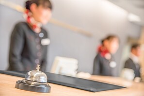 A hotel reception area with a wooden counter, a silver bell, and blurred figures of staff in the background.