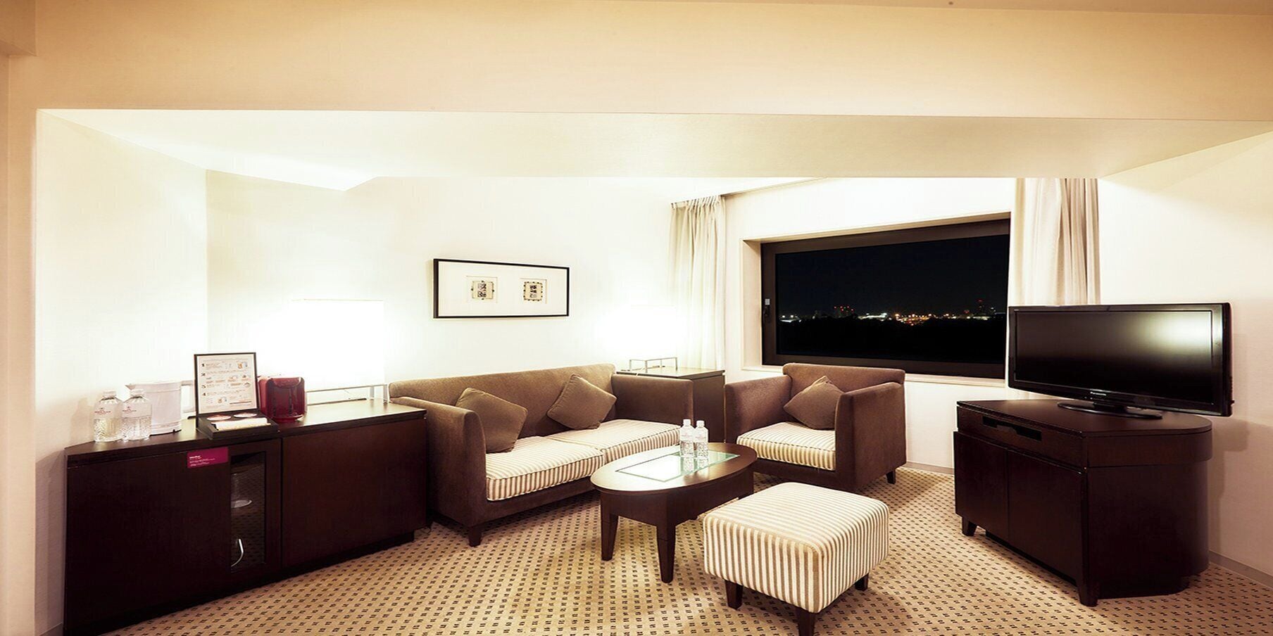 A hotel room with a sofa, armchair, ottoman, coffee table, TV, and a desk with a lamp and water bottles.