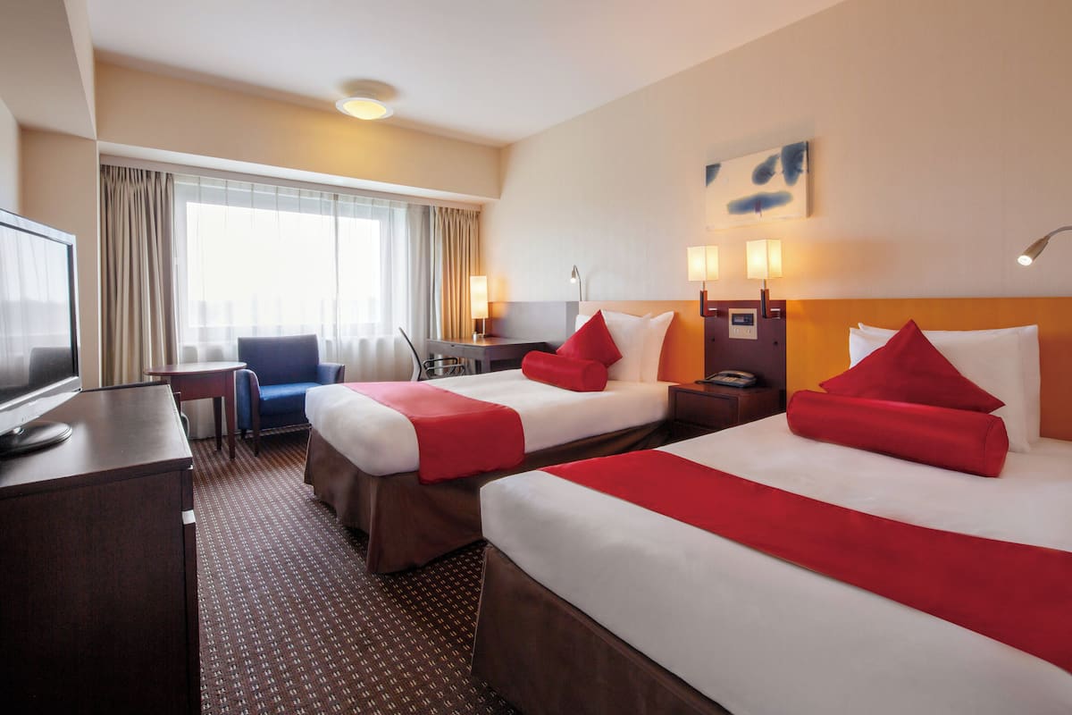 A hotel room with two beds, a desk, a chair, a TV, and a window with curtains.