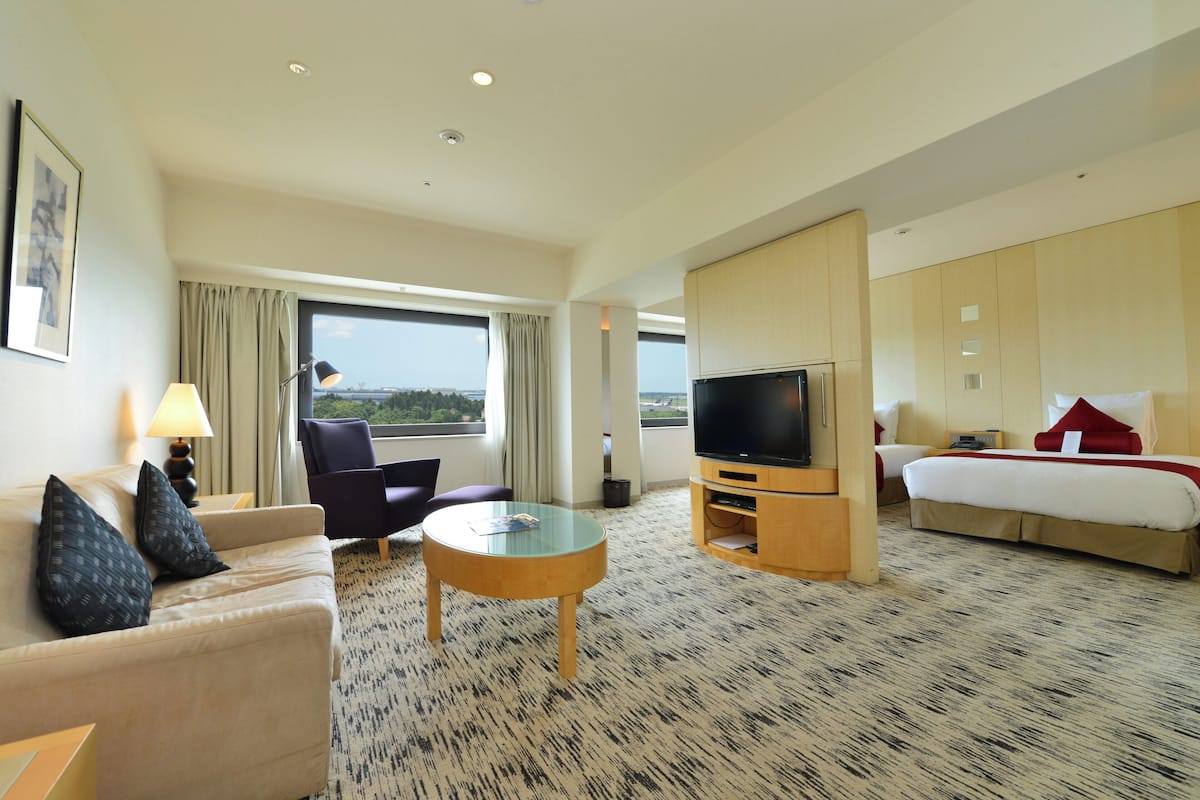 A hotel room with a sofa, armchair, TV, bed, and a view of the outdoors.