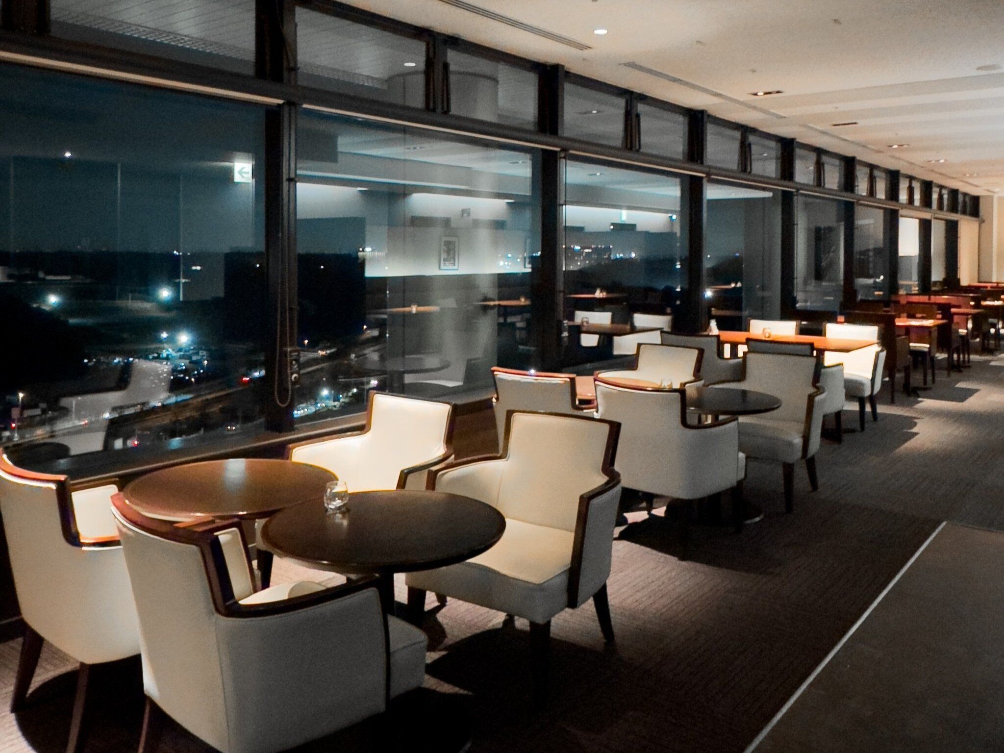 A modern indoor seating area with tables and chairs, large windows offering a view of the cityscape at night.