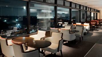 A modern indoor seating area with tables and chairs, large windows offering a view of the cityscape at night.
