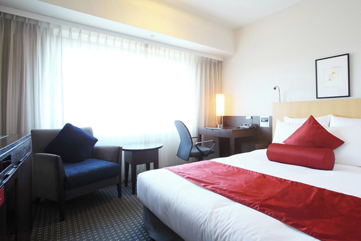 Een hotelkamer met een bed, een bureau, een stoel, een lamp en een raam met gordijnen.