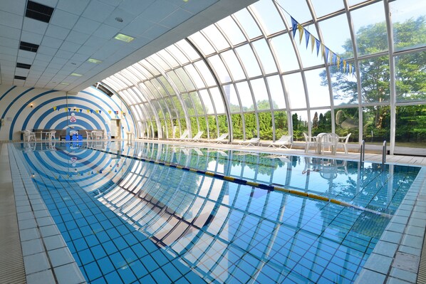 An indoor swimming pool with clear glass walls, lounge chairs, and a blue tiled floor.