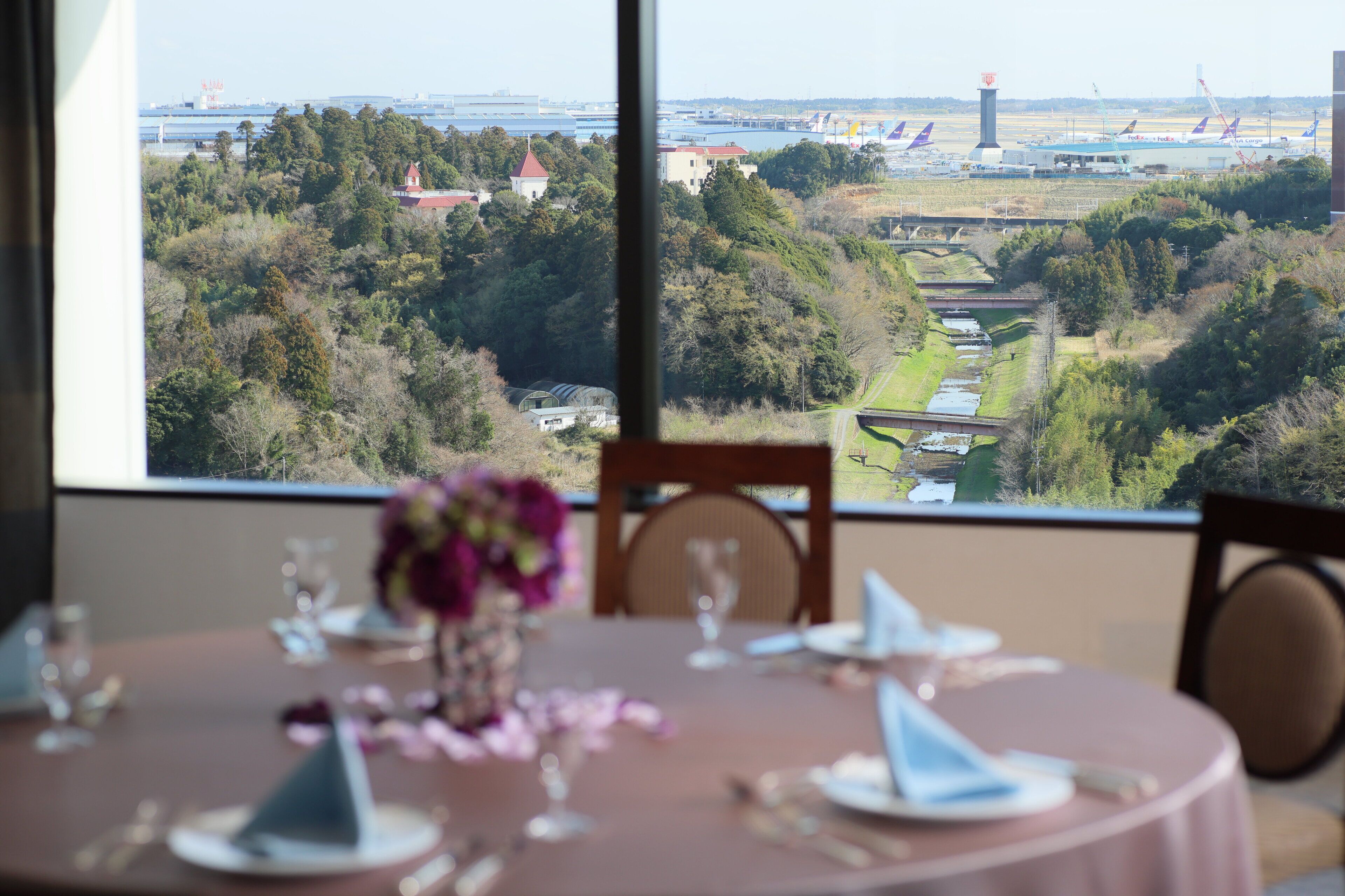 Uma mesa de jantar posta com um vaso de flores, vista de uma paisagem com árvores e um edifício ao longe.