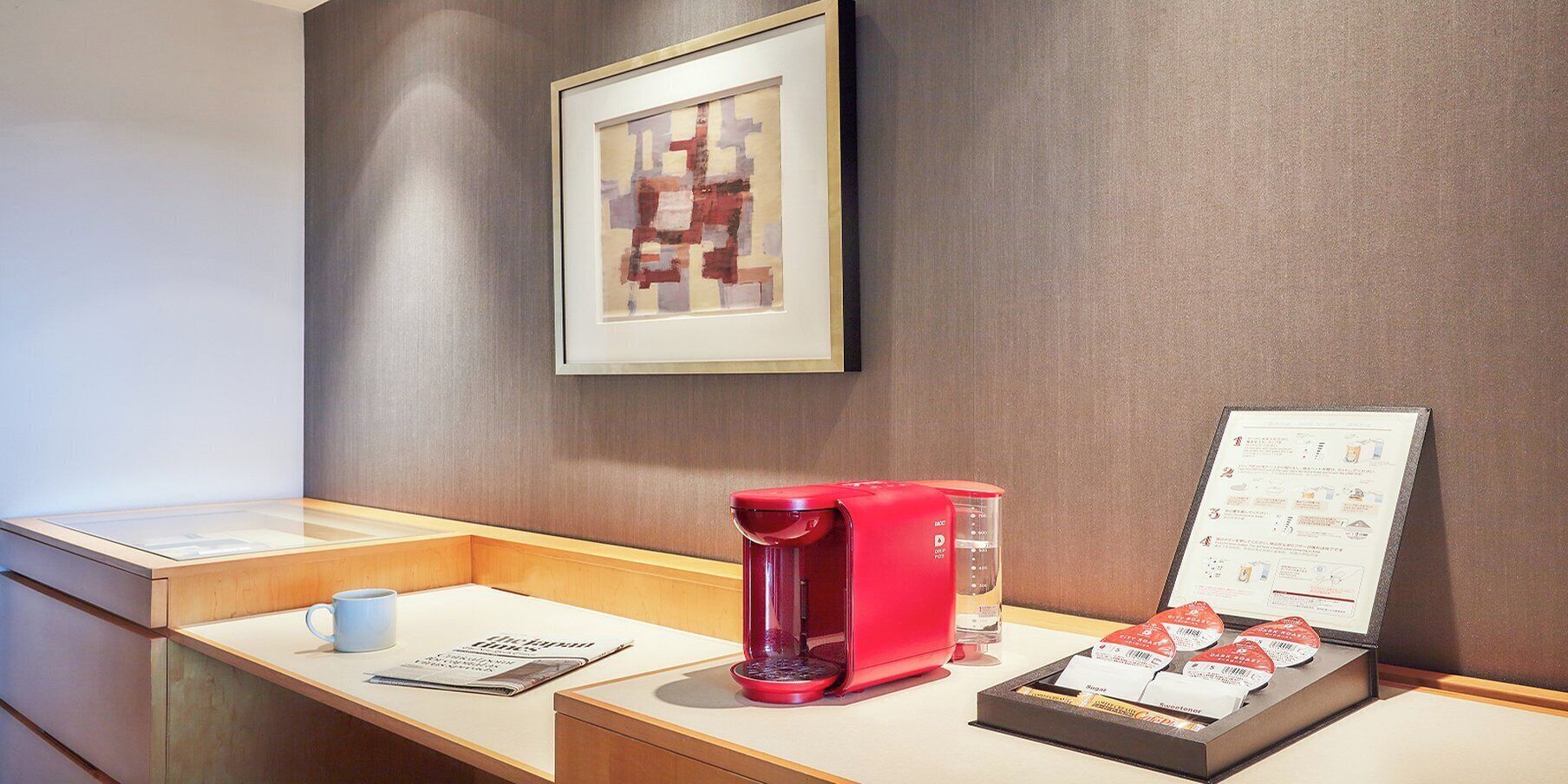 A hotel reception area with a framed abstract artwork, a red coffee machine, a menu stand, and a small table with a cup and a newspaper.