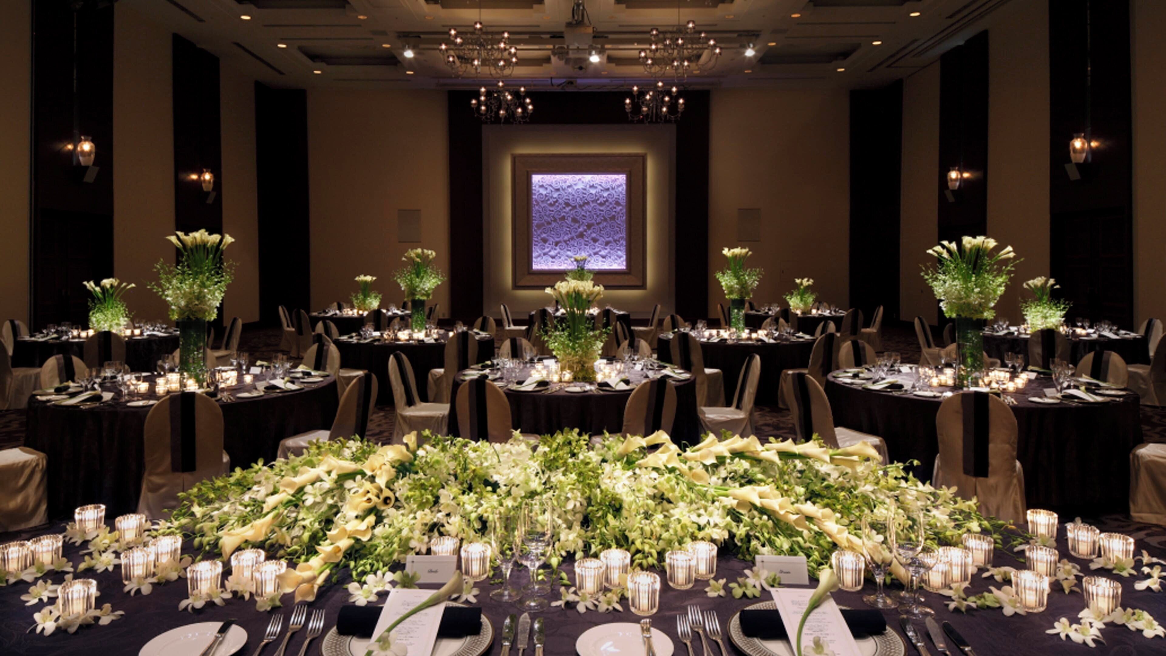 A banquet hall with round tables set for a formal event, featuring floral centerpieces and candlelit tables.