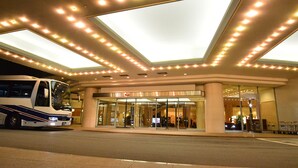Een hotelentree met een grote glazen gevel, een bus die ervoor geparkeerd staat, en een goed verlicht plafond.