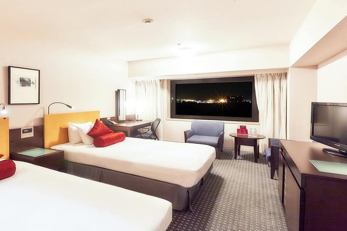 A hotel room with two beds, a desk, a chair, a television, and a window with a city view at night.